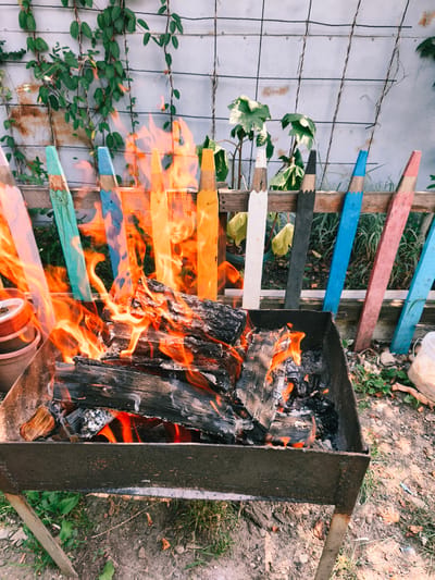 A fire burns in a metal brazier in an outdoor setting. The brazier is filled with burning logs and embers, with flames licking upwards. Behind the brazier, a wooden fence is decorated with colorful, oversized pencil-shaped posts. Vines climb on a wire fence in the background. The ground is covered with dirt, grass, and scattered leaves. In the foreground, to the left, a stack of terracotta pots is visible. The scene appears to be a backyard or garden area. There are no people or animals present in the image. The overall impression is one of a warm, rustic outdoor gathering or a preparation for a barbecue.