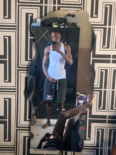 An indoor scene in Osara Gada, Nigeria, depicts a dark-skinned male subject captured in a full-length, irregularly shaped mirror selfie. The man stands barefoot, wearing a white tank top and dark gray shorts with "RHUDE" (reversed) and a small red and white bird logo on the left leg. He holds a black smartphone in his right hand, angled towards the mirror to take the photo, while his left hand is near the drawstring of his shorts.
The mirror reflects a dark wardrobe or cabinet behind the subject, upon which various items are placed: a black bag, several pairs of shoes (black and white sneakers), and a white tissue box labeled "T.C." To the right of the reflected wardrobe, a light brown wall is visible.
The wall surrounding the mirror is adorned with black and white geometric patterned wallpaper, featuring interlocking rectangular and L-shaped designs. At the bottom of the mirror's reflection, the man's legs are visible, along with a secondary, distorted reflection of someone holding a phone closer to the floor. The floor itself appears to be tiled. A small, purple, two-tiered plastic storage unit containing various items, and a small red object, are visible on the floor to the subject's right.
