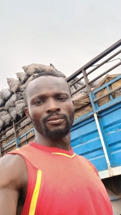 A man with a beard and a red t-shirt with a yellow stripe stands next to the open back of a truck loaded with sacks. The truck's blue sides and metal frame are visible. The sky is overcast. The man is the primary subject, positioned in the foreground, slightly off-center.