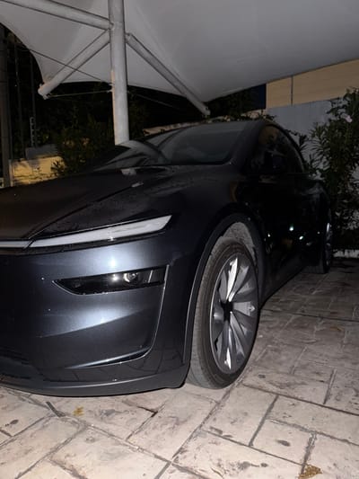 A dark gray Tesla Model 3 is parked on a stone patio under a white canopy structure. The car's front passenger-side wheel and part of the front bumper are visible. The car appears to be wet, possibly from rain, with water droplets on its surface. Green foliage and a building with yellow and blue accents are visible in the background. The scene is dimly lit, suggesting it is either evening or night. The image is taken from a low angle, focusing on the front of the vehicle. The patio is paved with rectangular stones. The location is identified as Mérida, Mexico.