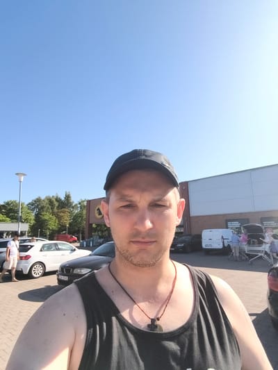In a self-taken photograph on a clear, sunny day, a man wearing a black baseball cap and a dark tank top stands in a parking lot. He is looking directly at the camera with a neutral expression. Around him, several cars are parked, and in the background, a modern building with a large yellow logo is visible. The building appears to be a retail store, possibly a supermarket, indicated by signage with text in German. One sign reads "Hier wieder ein Angebot – entdecken Sie deine persönlichen Eigenmarken mit der App." To the right, there is a shopping cart with people loading groceries into a car. Trees line the background, suggesting an outdoor, possibly suburban, setting. The bright sunlight and clear blue sky indicate it is daytime.