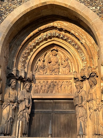 This image captures a highly detailed, weathered stone portal of a religious building, identified as a church or cathedral entrance in Brentwood, United Kingdom.

The scene is dominated by a deeply recessed archway, featuring multiple concentric archivolts adorned with intricate Gothic-style carvings of trefoils, quatrefoils, and foliage. The outermost arch is constructed from irregular, light-colored rough-hewn stone or flint.

At the apex, within the tympanum, a central relief depicts a seated, haloed male figure, presumably Christ, on a throne, with one hand raised. He is flanked by two kneeling, winged angelic figures, also haloed, facing inwards. Below this, a horizontal band presents a relief of approximately twelve to fourteen standing, robed figures arranged in a row.

Four standing, life-sized stone figures are positioned on pedestals within the door jambs. On the left, two bearded male figures in robes with halos stand, the inner figure holding a book. On the right, two similar figures are present; the inner, bearded figure holds a large book, and the outer figure, appearing younger, also holds a book or tablet.

Below the tympanum and between the jamb statues, a large, closed double wooden door, constructed from vertical planks, forms the entrance.

The stone carvings are illuminated by warm, directional sunlight from the upper left, casting deep shadows that accentuate the three-dimensional detail and give the stone a golden-brown hue. A dark metal finial is partially visible in the lower foreground.