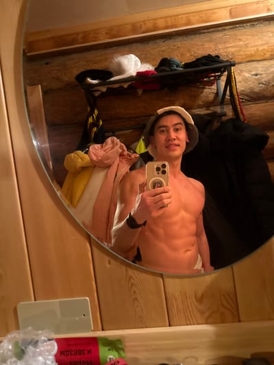 A man takes a selfie in a circular mirror. He is shirtless and wearing a white hat. His abs are visible. He's holding an iPhone. The background shows wooden walls and a clothes rack with various garments. A light switch and packaged product are visible to the side of the mirror. The image is slightly rotated.