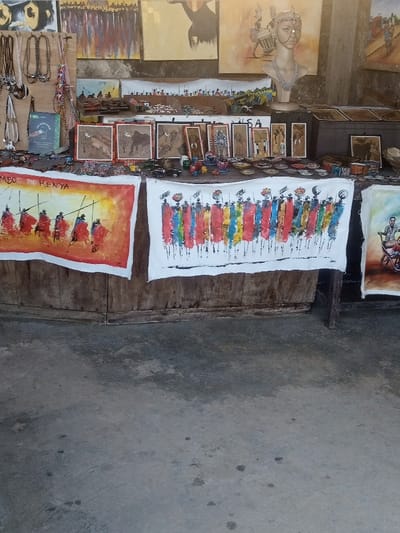 The image depicts an indoor or semi-outdoor art and craft stall, likely situated within a market area of Makadara/Mji Wa Kale ward, Mombasa, Kenya. The foreground consists of a grey concrete floor with various dark stains.
At the lower mid-ground, two large fabric or canvas paintings are prominently displayed on a wooden surface. The left painting features "JUMBO KENYA" text above a vibrant red and orange background with silhouettes of five figures, possibly Maasai warriors, holding spears and facing left. The central painting, on a white background, depicts a horizontal line of approximately twelve stylized figures in brightly colored, vertically striped traditional attire. A partial view of a third painting is visible to the right.
Above these large paintings, a tiered display table or shelf is filled with numerous smaller artworks and crafts. Several framed pieces portray African wildlife, such as an elephant and a zebra, alongside other abstract or figurative designs. Small decorative items, including miniature vehicles and animal figurines, are scattered across the display surface. A collection of small, round, flat decorative items (possibly coasters or dishes) is arranged centrally. A light-colored bust of a female head, adorned with a metallic necklace and headpiece, is positioned towards the upper right of this tier. A dark, chest-like object is visible to the right of the bust.
The background wall is extensively covered with various larger paintings, depicting landscapes, abstract figures, and wildlife. To the far left, a vertical wooden panel displays multiple necklaces and other hanging accessories. A green book or pamphlet, with "KENYA" visible on its cover, is also displayed on the left side. No individuals are visible in the scene.