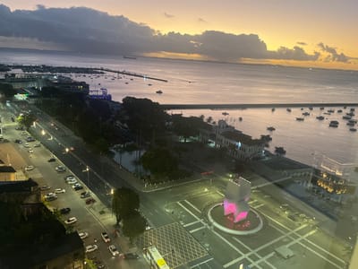 An elevated, wide-angle view captured at dusk depicts a vibrant urban waterfront scene in Salvador, Brazil, specifically overlooking Praça Cairu and Baía de Todos os Santos. The sky presents a gradient of color, transitioning from a warm orange-yellow near the western horizon to a cooler purple and dark grey at the top, indicative of heavy cloud cover.
In the foreground, an illuminated modern monument, composed of two large block-like structures, stands centrally in a circular fountain or reflecting pool, emitting a bright pink light. This monument, the Monumento ao Cruzeiro Quatrocentenário de Salvador, is situated within a large paved plaza marked with white road lines and yellow crosshatch patterns. A yellow bus is partially visible at the bottom edge of the frame. Reflections of interior lighting are visible on the glass surface through which the photograph was taken, primarily in the lower right quadrant.
Midground elements include a multi-lane roadway running parallel to the coastline, illuminated by streetlights, with several vehicles, some displaying active headlights or taillights. Numerous cars are parked along the road's edge. Adjacent to the road, historical buildings with white facades and prominent windows line the waterfront, including a structure resembling the Mercado Modelo. A small, tranquil body of water or canal is discernible behind a line of trees. Further along the waterfront, a distinct blue illuminated structure, possibly a stage or architectural feature, is visible.
The background is dominated by the expansive Baía de Todos os Santos. A long pier or breakwater extends into the bay, sheltering a significant number of small boats and vessels anchored within the calm waters. Beyond this sheltered area, the bay contains scattered larger ships, likely cargo vessels, in the distance. The far horizon shows the faint outline of land on the opposite side of the bay, dotted with distant lights.