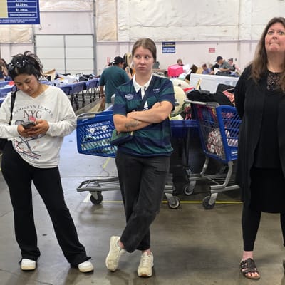Three individuals are depicted in a large, indoor retail or sorting facility, likely a Goodwill store in Milwaukie, Oregon. The woman on the left, of Latina descent, wears a white long-sleeved shirt with "Brooklyn League NYC" and "Tennis Club" graphics and black pants. She is looking down at her smartphone. In the center, a young Caucasian woman with light brown hair stands with her arms crossed, wearing a dark green and blue striped rugby-style shirt with a white collar and dark pants. She has a tattoo on her left arm and a small bag slung across her body. On the right, a Caucasian woman with long brown hair wears a black cardigan over a black top and black leggings, with dark sandals. She is looking upwards and to her left. The background reveals numerous blue shopping carts filled with merchandise, tables laden with items, and large white garage-style doors, suggesting a large volume of donated goods. Signage indicating prices like "$2.29 EA." and "$1.29 EA." is visible on a blue sign in the upper left corner. The floor is concrete, and the lighting appears to be overhead industrial lighting.