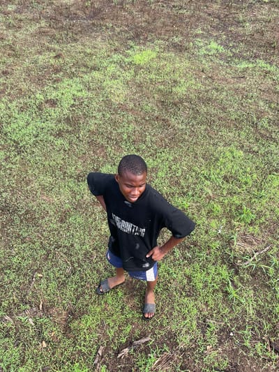 A young man stands in a grassy area, likely outdoors. He is viewed from above at an angle. The man is wearing a black t-shirt, blue shorts with a white stripe, and dark-colored sandals. The environment consists of varying shades of green grass with patches of bare earth. The location is Agba Umana, Nigeria.