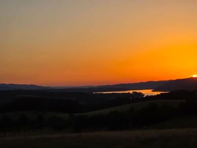 The image shows a sunset over a hilly landscape with a body of water. The sky is a gradient of orange and yellow, with the sun partially visible on the horizon. In the foreground, there are dark silhouettes of hills and trees. A body of water reflects the sunset colors, appearing as a bright orange stripe across the middle ground. Distant mountains are visible as dark blue silhouettes against the bright sky. The location is specified as Golyama Zhelyazna, Bulgaria. There are no people, actions, or interactions depicted in the image. The environment is natural, with rolling hills, trees, and a lake or reservoir. The time of day is sunset.