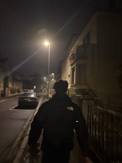A street at night illuminated by streetlights features parked cars and buildings along the sides. A person, wearing a dark jacket with a visible logo of "The North Face," is in the foreground, partially obscuring the view. The person is in motion, likely walking or riding a bicycle, as evidenced by the slight blur. The scene is dark and has a grainy texture due to low light conditions.