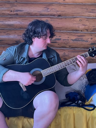 A person with dark, curly hair is playing an acoustic guitar. They are seated, and the guitar is black with a white pickguard and a light-colored fretboard. The background consists of a wooden wall with horizontal logs and rope detailing. The person is wearing a green jacket over a black t-shirt and dark shorts. They are positioned on a surface covered with yellow fabric, and to their right, there are some folded items, including a blue bag and a black leash. The scene appears to be indoors, possibly in a rustic setting like a cabin or dacha. The image does not provide any specific information about the location in the city of Трасна Снт, Russia.