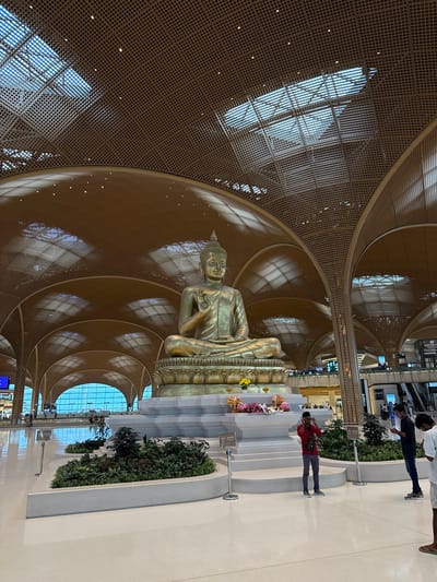 In an indoor space with a high, lattice-like ceiling, a large, golden statue of a seated Buddha is the central focus. The statue is positioned in front of a series of tiered platforms adorned with flowers. Sunlight streams in through large windows, creating bright patterns on the patterned ceiling.
Two individuals are visible in the scene. One person, wearing a red shirt and dark pants, stands near the platforms, looking towards the statue. Another person, wearing a white shirt and dark pants, is also present, though their activity is less clear. The overall atmosphere appears serene, with the grandeur of the statue and the bright, airy space suggesting a place of worship or reflection.
The setting appears to be a modern building, possibly an airport terminal or a large public space, given the expansive architecture and the presence of what look like shop signs in the background. The lighting suggests it is daytime.
The text visible in the image includes what appears to be directional signs or advertisements in the background, too small to be legible. The location context of "កណ្ដាលស្ទឹង, Cambodia" provides a geographical marker for the scene.