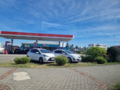 This is a photograph taken on a sunny day at a REWE Express gas station and convenience store in Walldorf, Germany. The gas station canopy, with its red trim and white underside, is a prominent feature, extending over several fuel pumps. Two cars are parked near the store: a white Toyota Yaris with a German license plate "MTK CZ 688" and a white Opel Corsa with a German license plate "GG PP 100". Behind them, a dark-colored car is parked near the pumps. The store itself has a blue facade with "REWE express" and "backfrisch" signage. In the background, there is a modern white building with orange accents and trees. The foreground consists of a paved area with interlocking gray bricks and a decorative strip of red bricks. The sky is blue with scattered white clouds. No people are visible in the image.