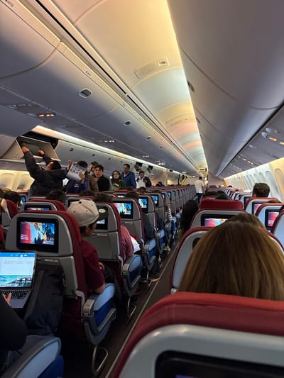 The image shows the interior of an airplane cabin. Passengers are seated in rows, some with screens displaying content on the seatbacks. Overhead bins are visible along the ceiling. One passenger is accessing a bin. Another passenger is working on a laptop. The location, Pudahuel, Chile is not visible or verifiable.