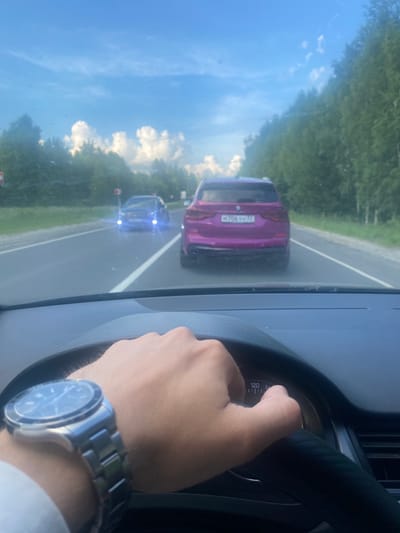 An interior shot from the driver's perspective of a car on a paved road with a slightly cloudy blue sky overhead. The driver's hands are visible on the steering wheel. The driver is wearing a wristwatch on their left wrist. The car's dashboard shows a speedometer with a needle pointing between 120 and 140. In front of the car, two other vehicles are on the road. The closest vehicle ahead is a purple SUV with a license plate that appears to read "M706YM 31". Further ahead, a blue sedan is visible with its headlights on. The road is flanked by trees on both sides. The scene is located on a road in or near the city of Beryozovka, Russia, suggested by the license plate format.