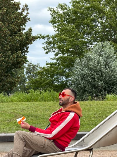 In Boxtel, Netherlands, a person wearing a red hooded jacket, tan cargo pants, and heart-shaped red sunglasses sits on a metal slide in a grassy park. The person is holding a small orange and white object in their right hand. The background features green trees and a cloudy sky.