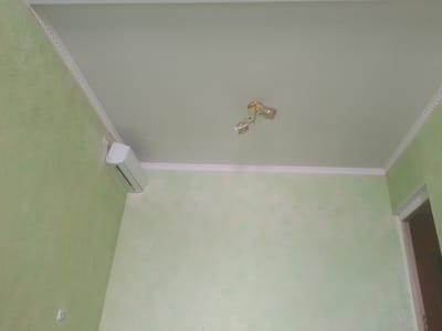 The image shows an interior view of a room, likely a living room or bedroom, with pale green walls and a white ceiling. A decorative white molding runs along the junction of the walls and ceiling.
Mounted on the wall near the corner is a white air conditioning unit. On the ceiling, a gold-colored light fixture with two glass shades is visible. The light fixture appears to be a ceiling spotlight or a similar type of decorative lighting.
The room appears to be sparsely furnished or empty, with no visible furniture or people. The lighting in the room is diffused, suggesting it might be daytime with indirect sunlight or artificial lighting.
The specific location within the city of Vahdat, Tajikistan cannot be determined from the visual information provided in the image. The image depicts a generic interior space that could be found in many residential or commercial buildings.