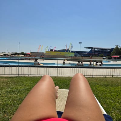 This is a first-person perspective, eye-level shot of a person lying on a lounge chair in what appears to be an outdoor swimming pool complex, likely during the day under a clear blue sky. The foreground is dominated by the person's legs, which are tanned, and the beige woven material of the lounge chair. A sliver of blue fabric, possibly part of swimwear, is visible at the bottom.
In the midground, a swimming pool with clear blue water is visible, separated by a white-tiled edge and a light-colored metal fence. Several people are in or around the pool. Some are standing, some are sitting, and some appear to be interacting with inflatable toys. A group of people are gathered near the far edge of the pool, some under umbrellas.
The background features more of the pool area, with buildings that suggest a sports or leisure facility. There are tall light poles and flags with various colors and logos, including what appears to be "Arena" written on some of them, suggesting a connection to sports or swimming. Further in the background, a large, multi-tiered grandstand or stadium structure is visible, indicating a larger complex. Trees are also present behind the buildings. The overall impression is a sunny day at a public pool or water park.
While the exact location in Reggio nell'Emilia, Italy, cannot be definitively determined from the visual information alone, the presence of the "Arena" branding and the architectural style of the grandstand might suggest a sports facility within or near the city.