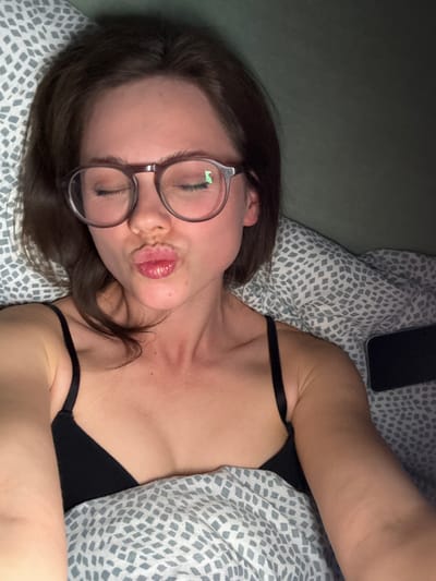 A woman with brown hair and round glasses lies on a bed with a gray and white patterned duvet. She is wearing a black tank top and making a kissing face towards the camera. A black smartphone rests on the pillow above her head. The background includes a pale green wall. The location cannot be confirmed as Kranj, Slovenia based on the visual information provided.