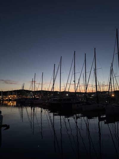 The image captures a marina in Lavreotiki Municipal Unit, Greece, at dusk. The sky transitions from a deep, dark blue at the top, where several distinct points of light (likely stars or planets) are visible, to an orange and pink gradient at the horizon, indicative of a recent sunset. Numerous sailboats are docked in the foreground and midground, their tall, slender masts forming dark, vertical silhouettes against the illuminated sky. The calm water surface reflects these masts as elongated, undulating lines. On the distant shore, buildings are visible, some with interior or exterior lights illuminating their facades and casting warm, bright reflections onto the water. Hills or landmasses form a dark, undulating line beneath the horizon. No individuals are visible within the frame.