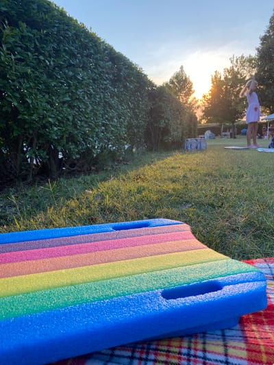 In Deutschkreutz, Austria, at sunset, a colorful floating mat rests on a red and white plaid blanket on a grassy lawn. The mat, featuring stripes of blue, purple, pink, orange, yellow, and green, has two handle cutouts. Behind the mat, a manicured green hedge runs horizontally across the frame, with tall grass and scattered weeds in the foreground. In the midground, a young girl in a white and pink striped dress stands facing away from the camera, her arm raised. Other people and various objects, including tents and mats, are visible in the distance on the lawn. The sun is setting, casting a warm glow and creating lens flares through the trees in the background, under a clear sky.