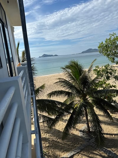 The image captures a view from a balcony overlooking a sandy beach and the ocean in Song Cau, Vietnam. A portion of a white building's exterior, including windows and a railing, occupies the left side of the frame. In the foreground, a palm tree with dark green fronds stands on the sand, partially obscuring the beach and the calm, blue-green ocean. In the distance, a low-lying island or peninsula is visible across the water under a sky filled with white, scattered clouds. The scene suggests a tropical coastal setting.