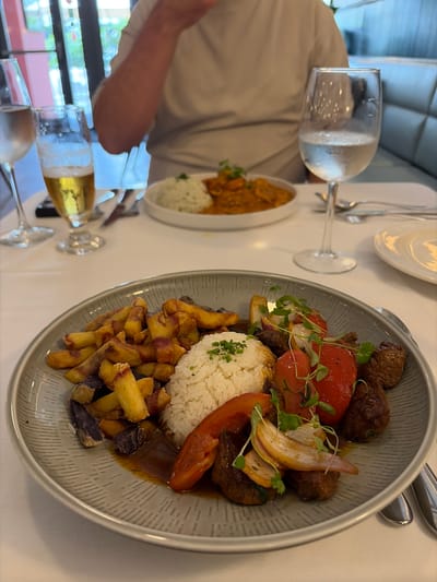 A high-angle shot captures a dining scene at a restaurant, potentially in Coral Gables, Florida, based on context clues (not definitively evident in the image). The composition is dominated by two plates of food and glassware on a light-colored tabletop, possibly a tablecloth.
The primary focus is a grey-rimmed plate containing a serving of Lomo Saltado, a Peruvian dish. The dish includes a mound of white rice topped with parsley or herbs, strips of sautéed beef, sliced red bell peppers, fried potatoes (some with purple skin), and onions. The plate is partially filled with a sauce or gravy.
To the left and slightly behind this plate is another smaller, white plate filled with a yellow curry-like dish and a scoop of white rice, presumably an additional menu item.
Two wine glasses are visible. The first glass contains a clear liquid, possibly water, with condensation on the outside. The second glass holds a golden liquid, likely beer or white wine, with visible bubbles. There are silverware pieces near the main plate.
In the lower left corner, a person’s arm and torso are visible, clad in a light-colored shirt. The background is indistinct, possibly showing a glimpse of a window with tree branches. The overall lighting suggests an indoor setting, possibly near a window, due to the natural light present in the scene.