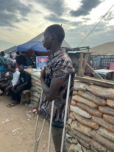 In Oko Amakom, Nigeria, a young man stands with the aid of crutches, his gaze directed to the left. He is wearing a patterned shirt and dark trousers. The background depicts a bustling marketplace scene with several other individuals seated and standing. A stall with a blue awning displays a sign indicating "POS" services with options for withdrawal, transfer, and deposit. Stacks of "BUA CEMENT" bags are prominently positioned to the right of the main subject, serving as a barrier. The environment appears to be an outdoor, unpaved area, with debris scattered on the ground. The sky overhead is cloudy.