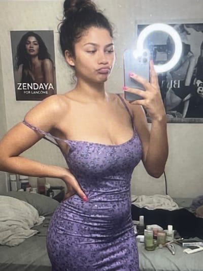 The image shows a young woman taking a selfie in what appears to be a bedroom. She is wearing a purple, patterned, fitted dress. Her hair is pulled up in a bun. She is holding a smartphone in her right hand and her left hand is on her hip. Behind her, a poster of Zendaya for Lancôme is visible on the wall, as is a ring light. The room appears somewhat cluttered, with a bed and various items on a surface that may be a desk or dresser. The background details are indistinct and do not provide any specific information about the location within Pruszcz Gdański, Poland.