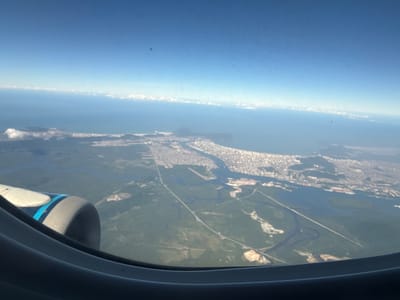 An aerial view from an airplane window captures a vibrant coastal urban landscape under a clear blue sky. In the bottom left, the dark grey frame of the aircraft window is visible, with a portion of a jet engine nacelle, featuring blue and white markings, protruding into the frame. The scene unfolds to reveal a dense urban area, characterized by numerous light-colored buildings, stretching along a prominent coastline. This urban expanse is immediately bordered by the vast expanse of the Atlantic Ocean, which transitions into a clear blue sky dotted with a thin band of distant cumulus clouds near the horizon.
Below and adjacent to the main urban core, a significant port complex is discernible, featuring extensive infrastructure, canals, and waterways that weave through the land. Inland from the coastal city and the port, large green areas, likely comprising wetlands, mangroves, and other vegetation, are crisscrossed by a network of roads and smaller rivers. Some lighter, cleared patches within these green areas suggest ongoing development or industrial activity.
Based on the distinctive coastal configuration, the sprawling port facilities, and the surrounding natural and developed areas, the image depicts the city of Santos, São Paulo, Brazil, with a particular focus on the Port of Santos and its immediate geographical context. No individuals or specific actions are discernible from this altitude.