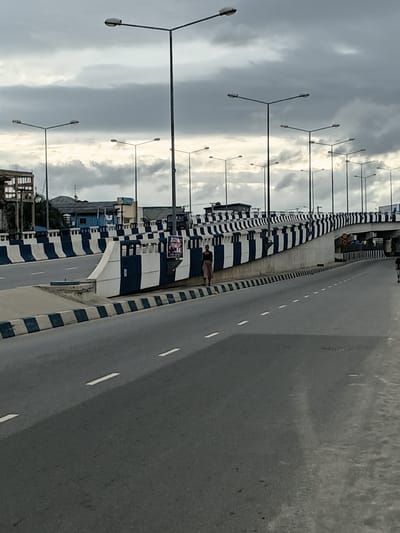 The image shows a highway overpass in Rumuokwuta, Nigeria, on a cloudy day. The overpass is painted with alternating black and white stripes. A few streetlights are visible, along with buildings in the background. There are at least two individuals on the side of the road. One person is standing near a guardrail, and another is walking further down the road. The road itself is paved and has white lane markings. The sky is overcast, suggesting a potentially humid or rainy climate.