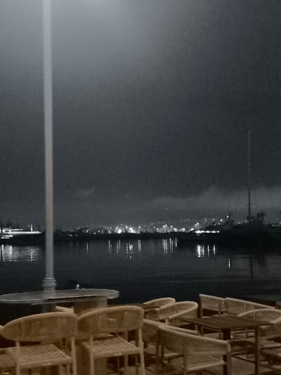 This nighttime photograph presents a tranquil waterfront scene in Palaio Faliro, Greece. The foreground features an outdoor seating area, equipped with several empty rattan-style chairs and tables, suggesting a quiet moment at a cafe or restaurant. A tall, light-colored pole stands prominently on the left side, extending vertically through the frame.

Beyond the seating area, a calm body of water stretches across the midground, serving as a reflective surface for the distant city lights. Across the water, the background reveals a cityscape with numerous twinkling lights, indicating a populated area nestled against a hill or shoreline. On the right side of the water, the tall mast of a sailboat is visible, silhouetted against the dark sky. On the left, a pier or dock area with some illumination can be seen.

The sky is dark and overcast with visible clouds, contributing to the atmospheric nighttime setting. No people or specific activities are depicted, giving the scene a serene and almost contemplative mood. The image has a desaturated aesthetic, dominated by dark blues and grays, with the chairs and tables offering a warm, muted brown tone. No visible text is present.