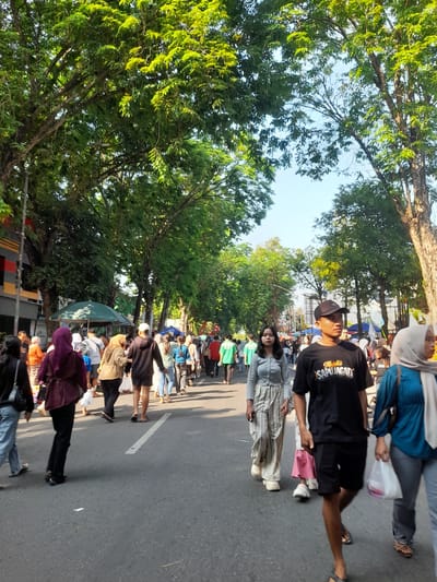In Purwodadi, Indonesia, a street is lined with mature trees casting dappled shade on a moderately busy scene. The paved road, marked with a white dashed line, is occupied by a diverse crowd of people walking in various directions.
In the foreground, a young man wearing a black t-shirt with "Majelis SAPUJAGAD" printed on it, black shorts, a black baseball cap, and sandals walks towards the viewer. To his left, a young woman in a light gray long-sleeved top and striped pants also walks towards the viewer. To the right of the foreground male, a woman with a light gray hijab and a teal top is visible.
Further back, a woman in a maroon hijab and black pants is walking away from the viewer. Other individuals are seen in the middle ground, some carrying shopping bags. There are several market stalls or vendor tents visible along the sides of the road, with green canopies. The buildings on the left side of the street appear to be commercial establishments. The sky is clear and bright blue, suggesting a sunny day. The overall atmosphere appears to be that of a lively street market or public gathering.