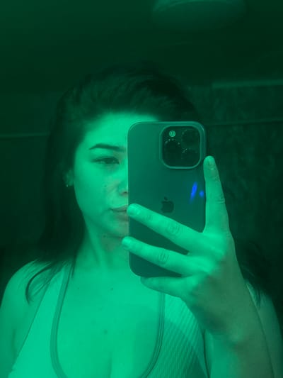 A woman takes a selfie in a mirror. She is holding a smartphone in her right hand and is wearing a light-colored sports bra. The image is lit with a green hue, giving the scene an eerie glow. The woman's face is partially visible, with her eyes looking towards the camera. The background is dark and appears to be a bathroom or dressing area. There are no distinguishing features to indicate a specific location within Gavrilov Posad, Russia.