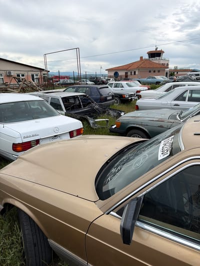 The image depicts an outdoor impound lot or junkyard in Natakhtari, Georgia, filled with a variety of older model Mercedes-Benz sedans. The vehicles are parked closely together on a grassy, unpaved surface. The sky is overcast and the ground appears damp, suggesting recent rain. In the background, a building with a distinctive orange-tiled roof and a control tower is visible, possibly indicating the site is near an airfield.

The foreground is dominated by the hood and fender of a gold-colored Mercedes-Benz, which shows signs of wear and tear, including water spots on the paint. A white sticker with black text and a barcode is affixed to the windshield of this car. To the left and slightly behind it is a white Mercedes-Benz sedan with "560 SEL" badging on the trunk. Further back, a dark blue Mercedes-Benz sedan is visible, as are several other similar vehicles in various states of disrepair, some with parts missing or bodies showing corrosion. A few other cars, including what appears to be a light blue sedan, are also present in the middle ground. No people are clearly visible or identifiable in the scene. The overall impression is one of a collection of abandoned or salvaged vehicles.