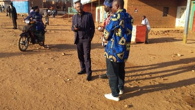 The image depicts a scene on a dusty street in Kayna, Democratic Republic of Congo. The street is made of loose dirt, with some scattered debris. A man in a black suit and tie is talking to another man wearing a blue patterned shirt, while a third man in a blue turban-like head covering stands behind them. They are standing in the middle of the street. A woman in a blue dress sits on a motorcycle to the left of the men. Behind them, a man wearing a white shirt stands by a red wooden structure. In the background, there are some buildings and other people.  The buildings appear to be made of brick or concrete, with some having metal roofs. The image is taken during the day, with the sun casting long shadows on the ground.