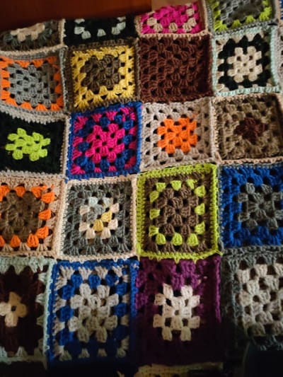 A crocheted blanket is spread out on a surface, possibly a bed or floor, in Rechnaya Sosnovka, Russia. The blanket is composed of many individual granny squares in a variety of colors including pink, orange, yellow, green, blue, brown, white, and gray. The squares are arranged in a grid pattern. The lighting suggests an indoor setting. There are no people or animals visible in the image. The focus is entirely on the texture and colors of the handmade blanket.