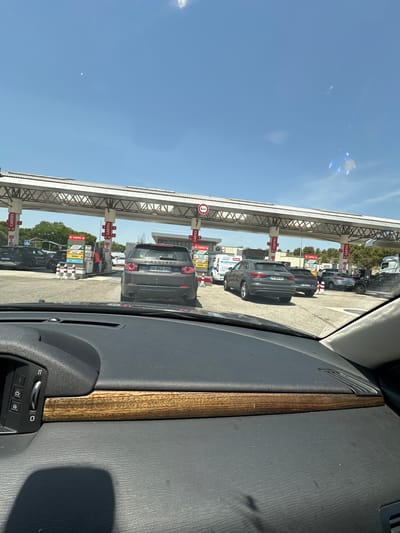 This is a photograph taken from inside a vehicle, looking out at a line of cars at a gas station. The cars are lined up under a covered structure with gas pumps. Several signs are visible, some with red lettering reading "CARTES" (French for "cards") and others indicating fuel types like "DIESEL" and "GPLC" (LPG). One car in the foreground is a dark gray SUV with the license plate "GQ-952-PR". Further down the line, another car has the license plate "FV-554-6S". The sky is clear and blue, suggesting a sunny day. The time of day appears to be midday, given the bright sunlight and shadows. The overall scene depicts a typical situation at a busy gas station, possibly on a highway rest stop given the covered pumps and orderly lines of vehicles. The perspective from inside the car, with a portion of the car's interior visible in the foreground, adds a personal viewpoint to the scene. The image has been rotated 90 degrees counter-clockwise for display.