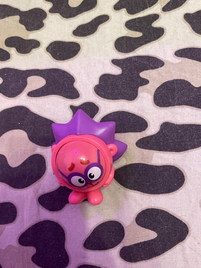A pink toy character with purple spiky hair and glasses is positioned on a fabric with a camouflage pattern. The toy has a round head, large white eyes with black pupils, and two small pink legs. The fabric features irregular dark gray and light beige splotches. The lighting in the image appears to be influenced by a purple light source, casting a pinkish hue on the toy and parts of the fabric. There is no discernible indication of a location in Borisova Griva Massiv, Russia.