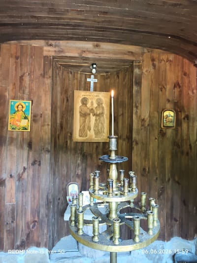 The image depicts the interior of a small, rustic wooden chapel or shrine located in Beli Osam, Bulgaria. The walls and ceiling are constructed from unpainted, light brown wooden planks, exhibiting visible grain and knots. The ceiling is slightly curved. In the central foreground, a multi-tiered, ornate brass or bronze candelabra stands on a stone base. A single, tall white candle with a visible flame is lit in the topmost holder of the candelabra; other candle holders on the lower tiers are empty or contain wax residue.
Affixed to the left wooden wall is a rectangular icon of Jesus Christ with a halo, holding an open book, rendered in vibrant colors. On the central back wall, within a recessed wooden frame, a larger, vertically oriented wooden icon or relief carving is displayed. This icon depicts two robed figures, identified by Cyrillic text above them as "СВ. АП. ПЕТЪР И ПАВЕЛ" (SS. AP. PETER AND PAUL), holding an object between them. Above this central icon, a small white cross is mounted on the wall, and above the cross, a light fixture with a bulb is visible. To the right, on the wooden wall, a smaller, dark-framed icon shows a figure riding a horse. The floor beneath the candelabra is a rough, light-colored stone or concrete. The timestamp "06.06.2026 15:59" and device "REDMI Note 15 Pro+ 5G" are present at the bottom of the image.