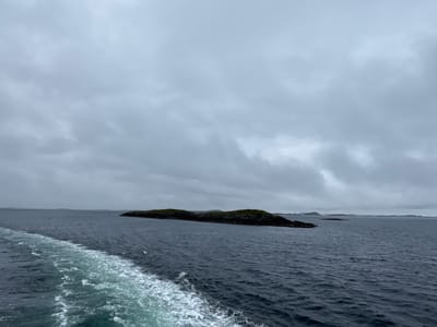The image captures a wide, overcast view of the ocean near Onøya, Norway. A ship's wake, a trail of white foam, cuts diagonally across the lower left portion of the frame, indicating movement. In the middle ground, a long, low-lying island covered in dark green vegetation stretches horizontally across the scene. The water is a deep, murky blue-grey, reflecting the dull, cloudy sky. In the distance, the horizon is dotted with faint, blurred outlines of other islands or landmasses. The overall atmosphere is somber and muted due to the overcast conditions. There are no people or specific buildings visible in the frame, only the natural elements of sea, sky, and islands.