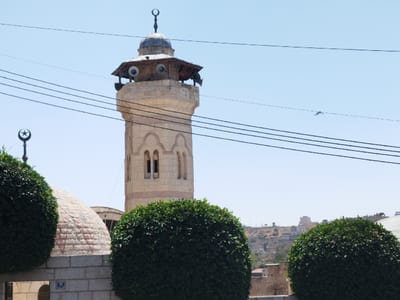 The image displays a daytime scene in Al-Khalil, Palestinian Territory, featuring a prominent minaret. A tall, light-colored stone minaret stands centrally, characterized by a multi-sided shaft with two arched windows visible on its facade. The minaret is crowned by a balcony featuring a wooden canopy, from which multiple white loudspeakers are mounted and project outwards. A dark, domed cap with a crescent finial tops the structure. In the lower-left foreground, a section of a light-colored stone dome is visible, surmounted by a metal finial shaped like a crescent and star. Two large, spherically trimmed green bushes occupy the immediate foreground, partially obscuring the bases of the architectural elements. Beneath the dome and bushes, a light-colored stone wall is present, bearing a small rectangular plaque with the number '28'. Several parallel black overhead utility wires traverse horizontally across the upper portion of the frame. The distant background reveals an arid, treeless hill dotted with various light-colored urban buildings. A small, dark, indistinct object is discernible in the upper right sky above the wires. The scene is brightly lit under a clear blue sky.