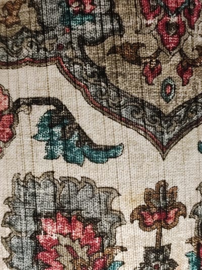 The image displays a close-up, textured view of a woven fabric with a floral and paisley-like pattern. The dominant colors are beige, cream, red, brown, and teal, with black outlines defining the motifs. The texture suggests a coarse weave, possibly from a rug, tapestry, or upholstery. No elements in the image provide any information about a specific location, including Roranwala, Pakistan, or details about people, actions, or interactions.