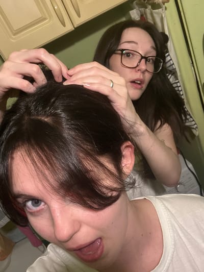 The image depicts two young women indoors. The woman in the foreground is looking directly at the camera with her mouth open, her eyes wide, and her head tilted. She has dark hair with some red highlights visible. The woman in the background is wearing glasses and has her hands in the hair of the woman in the foreground, seemingly styling it. She is looking towards the left of the frame with a surprised expression. The environment appears to be a domestic setting, possibly a kitchen or bathroom, with cabinetry visible in the background and a washing machine. The lighting is dim and artificial. There is no indication of the location in Obninsk, Russia.