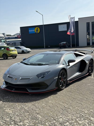 A matte grey Lamborghini Aventador SVJ is parked on a light grey interlocking paver surface in Meuspath, Germany, under a clear, bright sky. The vehicle is angled towards the right, presenting its front and left side. It features red accents on the front splitter and side skirts, with black multi-spoke wheels containing red center caps, and a prominent rear wing. To the left and behind the Lamborghini, a light green Ford Fiesta and a white Volkswagen Golf R are parked. Further in the background, a large, dark grey industrial building displays a prominent blue and yellow "BILSTEIN" sign with a shock absorber logo, and two vertical flags with "MANTHEY" branding. A streetlamp stands to the left of the Bilstein sign. To the far left, a partial view of a white truck and a sign for "DIENST MANHELLER" are visible. No individuals are present in the frame.