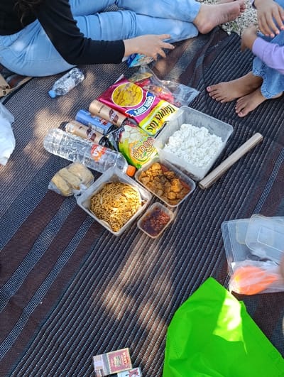 A picnic blanket lies on the ground, covered in a black and white striped fabric. It is laid out on a grassy surface in Kecamatan Banyuputih, Indonesia, located at coordinates -7.754 latitude and 114.367 longitude. There are several items laid out on the blanket, including: a white container filled with white rice, a red and yellow bag containing snacks, a container of fried noodles, a container of meat, a container of a dark red sauce, a box of matches, an orange bag, a green tablecloth, two plastic water bottles, and three cans of a dark beverage. On the left side of the image, a pair of legs clad in blue jeans can be seen. They belong to a person who is wearing a black top and sitting on the blanket. The person's left hand rests on the blanket near a red and yellow bag of chips. On the right side of the image, a pair of legs clad in blue jeans can be seen, belonging to a person sitting on the blanket. The person's right hand rests on the blanket near an orange bag. There are two people seated on the picnic blanket. The scene appears to depict a casual gathering. The environment is peaceful and relaxed.