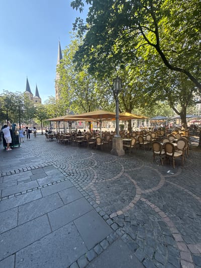 This is a daytime, eye-level, wide-angle photograph of the exterior seating area of a cafe in Bonn, Germany, with the Bonn Minster (Münster) visible in the background. The foreground is paved with large, rectangular grey stones, transitioning to a circular pattern of dark cobblestones. Numerous cafe tables and chairs, many with woven cane seats and backs, are arranged under large, beige umbrellas. Some tables are set, while others are empty. In the mid-ground, several people are walking and interacting. To the left, a person wearing a white shirt and shorts is pushing a green wheeled cart. Behind them, a few individuals are seen walking. In the background, the spires of the Bonn Minster rise above the trees. The sky is clear and blue, and the scene is bathed in natural sunlight, casting shadows from the trees and umbrellas.