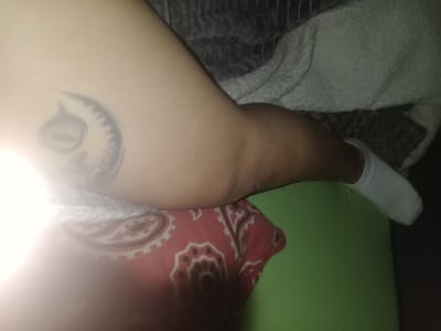 A person's leg is visible, lying on a bed or sofa. The person is wearing a white sock on their left foot. A tattoo is visible on the right thigh. The leg is resting against a red patterned pillow and a light green pillow. A white blanket is beneath the leg. The location is unknown, but the query mentioned Villa Ballester Noroeste, Argentina.