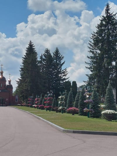 The image depicts a serene outdoor scene at a religious establishment, likely a church or monastery, in Kovalivka, Ukraine. The foreground features a paved pathway curving to the left, bordered by a manicured lawn. A line of lush green evergreen trees, including tall spruces and smaller firs, runs along the edge of the lawn. Interspersed among the trees are decorative flower beds filled with a variety of colorful blossoms in shades of pink, red, and white, some in urns and some planted directly in the ground.

In the background, a traditional red brick building with golden domes and crosses stands prominently on the left side of the frame. The building's architecture suggests a religious or historical significance. The sky above is a vibrant blue, adorned with fluffy white cumulus clouds, indicative of fair weather. The overall impression is one of peacefulness and natural beauty, enhanced by the careful landscaping and the presence of the religious structure. No people are visible in the image.