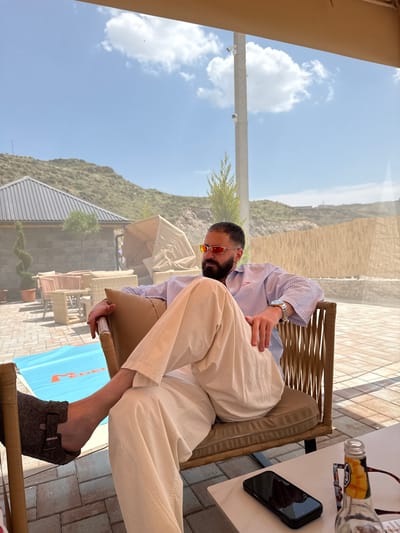 A man with a beard, wearing sunglasses, a blue and white striped shirt, and off-white pants, is sitting in a wicker chair with a tan cushion. He is wearing dark brown Birkenstock-style sandals. His left leg is crossed over his right. The chair is placed on a brick patio. In the background, there are several pieces of outdoor furniture, including wicker sofas and chairs, and a table with a blue mat on it. A building with a dark metal roof and stone walls is visible behind the furniture, as well as a hillside with sparse vegetation. A tall, thin pole with two security cameras is also present. The sky is blue with scattered white clouds. A bottle and a smartphone are on a white table in the foreground. The scene appears to be an outdoor seating area at a restaurant or cafe in Ptghni, Armenia, given the arid landscape and architectural style.