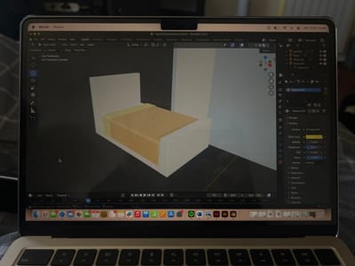 The scene depicts an interior located in Pedrouços, Portugal, focusing on an open silver Apple MacBook laptop. The laptop rests on a dark, textile surface, possibly a blanket or bed. Its screen displays the Blender 3D modeling software, version 3.5.1, with an active project file titled "quartoisometrico.blend". The primary viewport shows a simple 3D isometric model of a bedroom, featuring a white bed frame with a light orange mattress and bedding, positioned against a light gray wall and adjacent to a white vertical panel or wardrobe element. The macOS menu bar at the top indicates the date and time as "sáb. 21/02 12:36". The macOS Dock is visible at the bottom of the screen, displaying various application icons, including Spotify, Adobe InDesign, and Adobe Illustrator, with one icon showing a red notification badge containing the number "21". A partial view of the laptop's keyboard occupies the foreground. No individuals are present in the image.